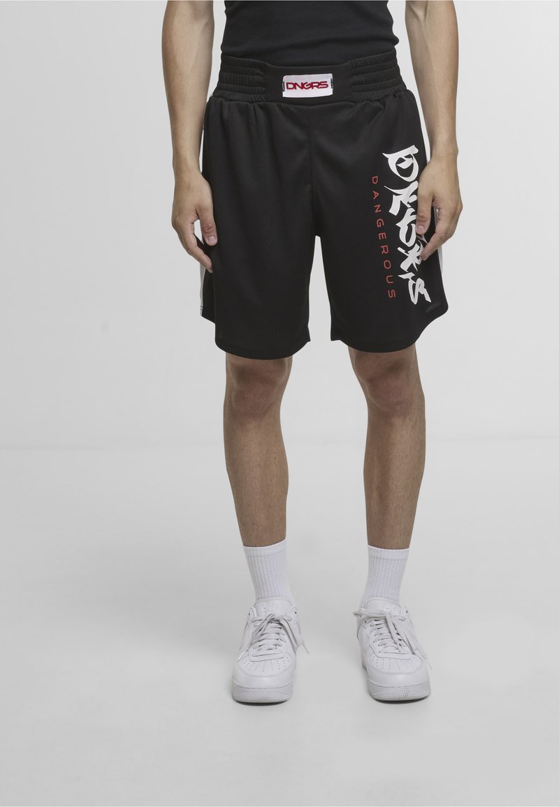 Personne portant un short noir avec le texte "DANGERS", des chaussettes blanches et des baskets blanches sur un fond clair uni.