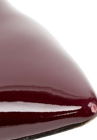 Gros plan sur un bout de chaussure pointu brillant de couleur bordeaux foncé avec une surface réfléchissante sur fond blanc.