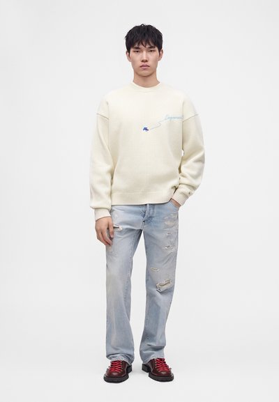 Jeune homme portant un pull crème avec une broderie bleue, un jean clair déchiré et des bottes foncées avec des lacets rouges, debout devant un fond blanc.