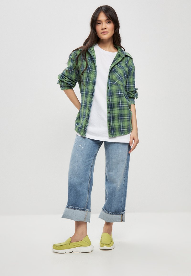 Cămașă flanelată în carouri verzi cu manșete suflate, purtată peste un tricou alb. Blugi albaștri deschis cu picior larg, cu margini suflate, și pantofi verde slip-on.