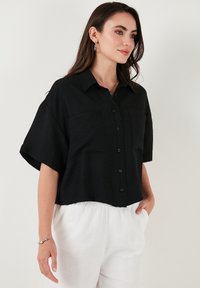 Camisetta nera a maniche corte realizzata in un tessuto testurizzato, con colletto, chiusura frontale con bottoni e due tasche sul petto.