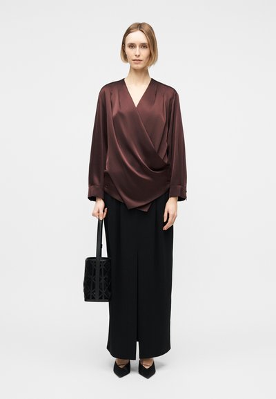 Γυναίκα που φοράει μπορντό σατέν πουκάμισο με δέσιμο, μαύρη μακριά φούστα με σχίσιμο μπροστά, μαύρα μυτερά παπούτσια και κρατάει μια μαύρη τσάντα χειρός.