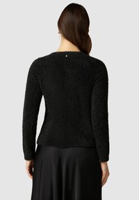 Svart texturerad tröja med långa ärmar och rund hals, med en knappdetalj bak och en något utsvängd nederkant.