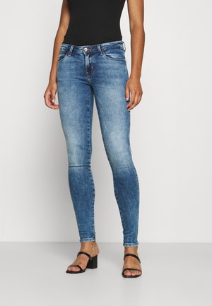 Femme portant un jean skinny bleu délavé, un haut noir sans manches et des sandales noires à talons avec brides, debout devant un fond uni.