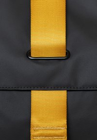 Borsa in tessuto nero con una distintiva tracolla a righe gialle e un anello metallico nero circolare per il fissaggio.