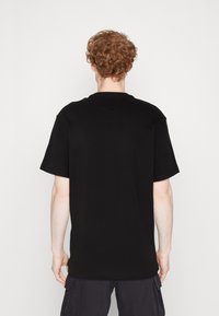 Man med lockigt rött hår sedd bakifrån, klädd i en enkel svart t-shirt med kort ärm och mörka cargo-shorts mot en vit bakgrund.