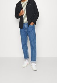 Chaqueta negra con cremallera, camiseta a rayas verdes y blancas, vaqueros azules holgados y zapatillas blancas con acentos rojos. Conjunto casual con un ajuste moderno.