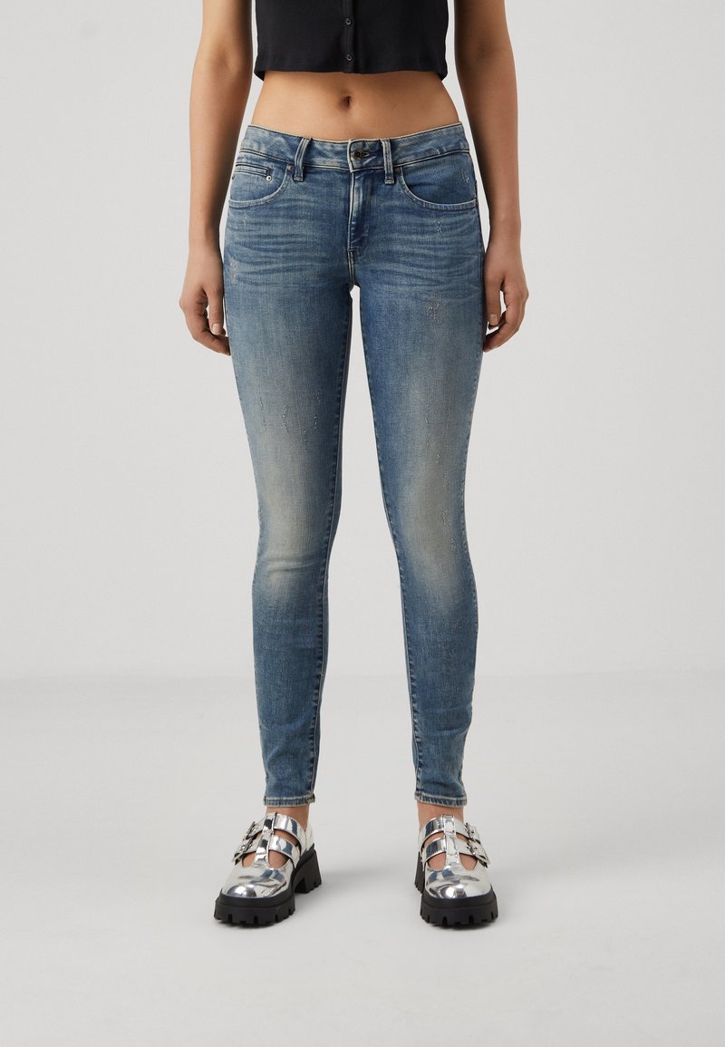 Jeans ajustados de denim azul claro con un ligero desgaste, con bolsillos frontales y cierre de botón plateado, combinados con zapatos metálicos.