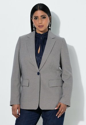 Frau trägt einen grau karierten Blazer über einer marineblauen Bluse mit Rüschenkragen und dunklen Jeans, steht vor einer weißen Wand.