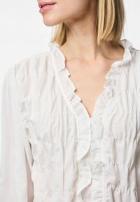 Blusa branca com decote franzido e tecido texturizado com costura horizontal. Mangas longas com um design leve e fluido.