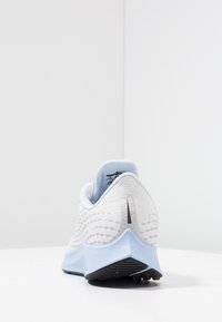 Scarpa sportiva bianca e azzurra chiara con tomaia in rete pattern, tacco arrotondato e suola in gomma. Presenta una striscia grigia di accentuazione sul tallone.