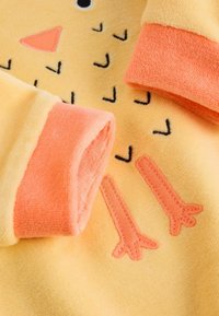 Gelbes Fleece-Gewebe mit orangen Bündchen. Mit einem Küken-Design, das einen rosa Schnabel, schwarze Augen und gestickte orange Füße zeigt. Weiche Textur.