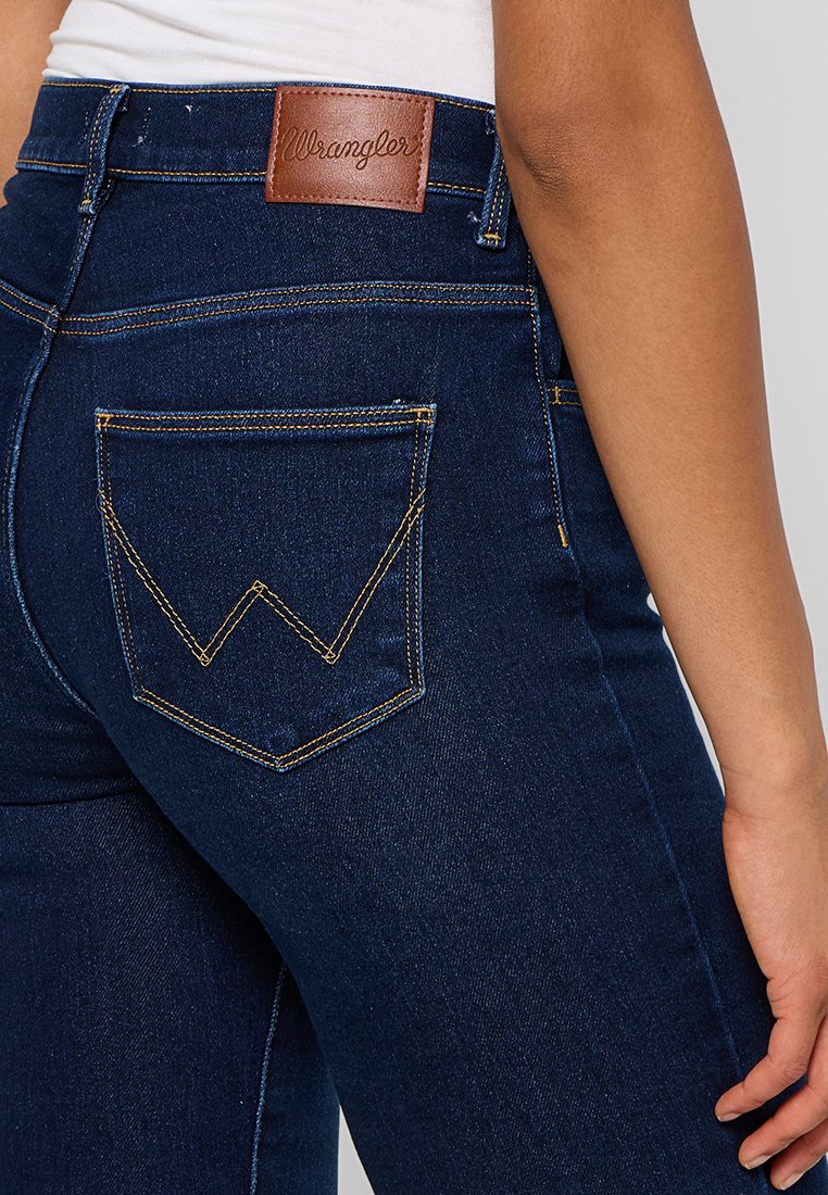 Jean en denim bleu foncé avec une taille haute, arborant un patch logo en cuir marron et des poches distinctives avec un design en chevron.