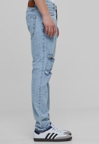 Jean en denim bleu clair avec une coupe slim, détails déchirés au niveau des genoux et une taille avec une pièce en cuir marron. Portés avec des baskets blanches et grises.
