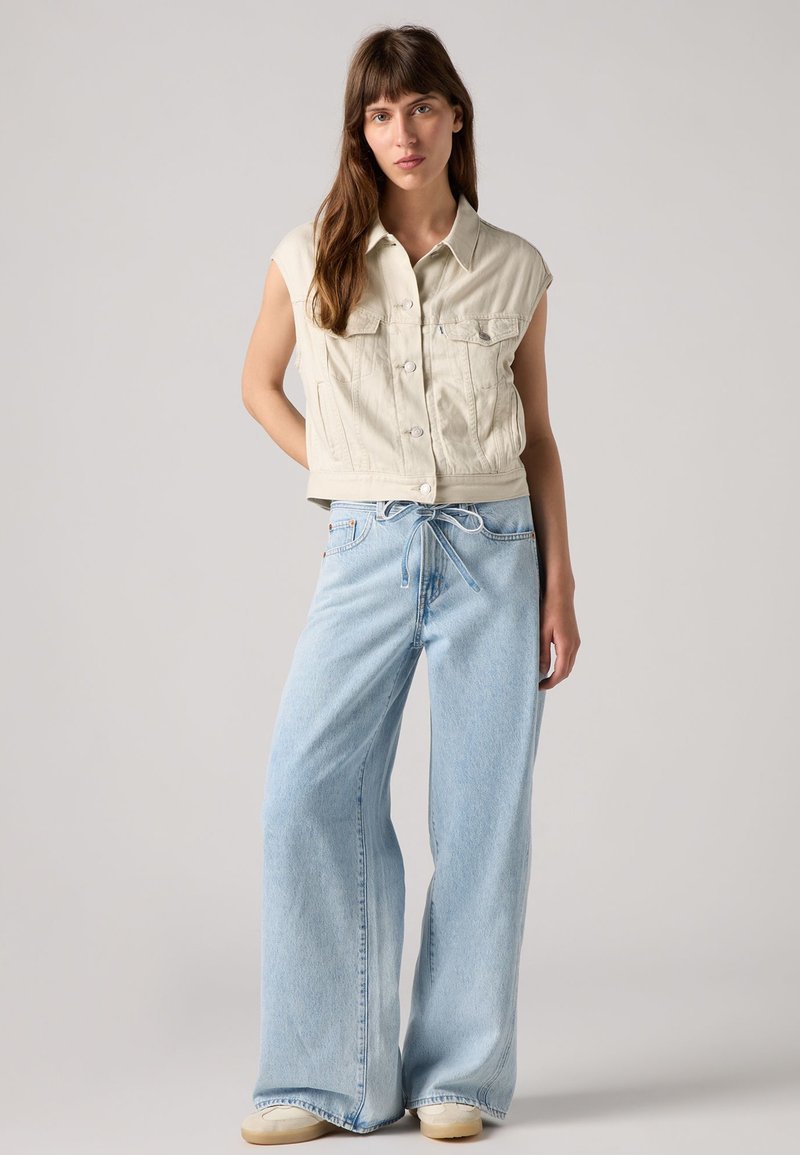 Gilet en denim crème coupé avec des boutons et deux poches poitrine, associé à un pantalon en jean bleu clair à jambes larges avec une taille à cordon et des baskets crèmes.