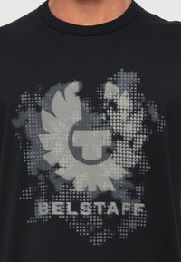 Črna bombažna majica z teksturirano sivo grafiko, ki vsebuje krožni logo in besedo "BELSTAFF" v debelih črkah na dnu.