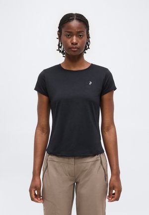 Kvinde med flettet hår iført en sort t-shirt med korte ærmer og beige bukser, stående foran en ensartet lys grå baggrund.