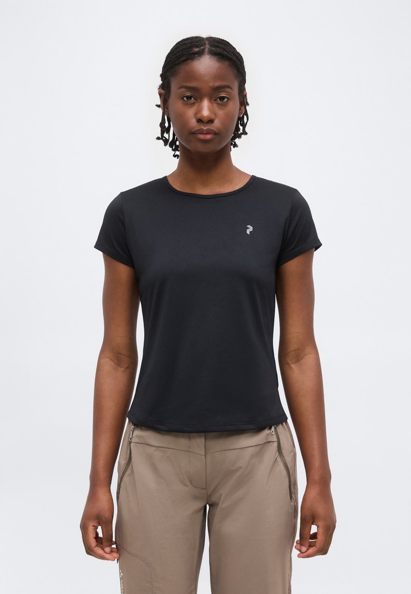 Γυναίκα με πλεξούδες στα μαλλιά που φοράει μαύρο κοντομάνικο μπλουζάκι και μπεζ παντελόνι, στέκεται μπροστά σε ένα απλό ανοιχτό γκρι φόντο.