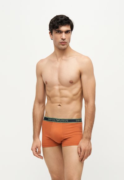 Homme musclé aux cheveux foncés portant un boxer orange Emporio Armani, debout devant un fond blanc uni.