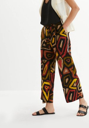 Persona che indossa pantaloni larghi con motivo geometrico astratto colorato, top nero senza maniche, giacca bianca senza maniche e sandali neri.