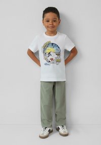 Witte T-shirt met een kleurrafische afbeelding van bergen en palmbladeren, gecombineerd met lichtgroene broek en witte sneakers met zwarte accenten.