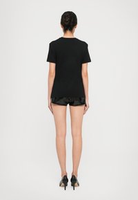 Woman standing with back visible, wearing a black short-sleeve shirt, shiny black shorts, and black high-heeled shoes against a plain background.