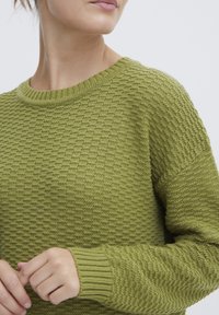 Grön stickad tröja med ett texturerat mönster, ribbad halsringning och mudd. Visar en närbild av tygets intrikata väv och färg.