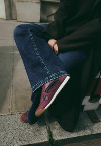 Maroon nahkahogot mustilla yksityiskohdalla, valkoisella pohjalla ja tekstuurilla ommellulla yksityiskohdalla; yhdistettynä sinisiin leveälahkeisiin farkkuihin, istuen kivisellä pinnalla.