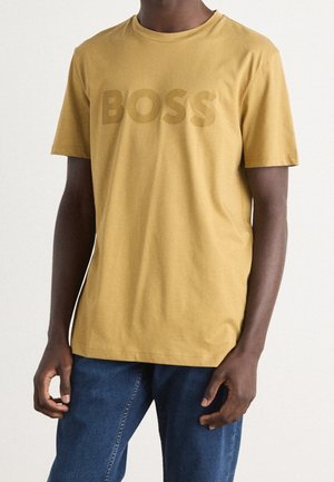 Mężczyzna w musztardowożółtej koszulce z krótkim rękawem z logo "BOSS" i niebieskich dżinsach, stojący na jednolitym jasnym tle.