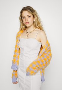 Cardigan de malha grossa com padrão laranja e lavanda, usado sobre um vestido branco justo com alças finas. O cardigan tem fio texturizado.