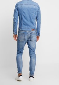 Man in een lichtblauwe spijkerjack en jeans, staand met ontspannen handen naast zich, gecombineerd met witte sneakers, van achteren weergegeven.