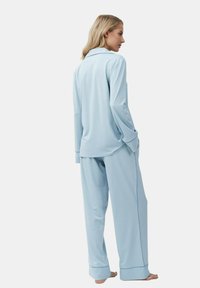 Femme debout pieds nus portant un top de pyjama à manches longues bleu clair et un pantalon de pyjama large assorti avec des détails de passepoil foncé.