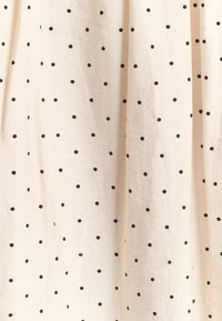 Jasno beżowa tkanina o teksturowanej powierzchni, pokryta równomiernie rozmieszczonymi czarnymi kropkami w różnych rozmiarach na całej powierzchni.