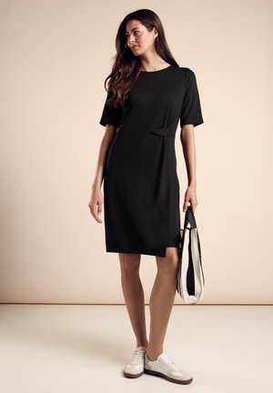 Zwart kort mouwen jurk met een cinched taille, knielange zoom en zachte textuur. Gecombineerd met een gestreepte tas en witte sneakers.