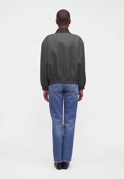 Personne debout de dos portant une veste gris foncé avec un col noir, un jean bleu et des chaussures noires sur un fond blanc uni.