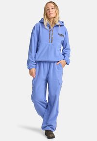 Ensemble en polaire composé d'un hoodie et d'un pantalon de couleur bleu clair. Le hoodie présente un cordon de serrage et des coutures décoratives. Le pantalon est doté de poches cargo et de poignets élastiques.