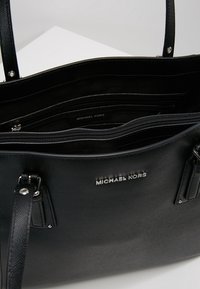 Zwarte leren tote bag met een gestructureerd oppervlak, zilverkleurige hardware, bovenhandvatten en een ruime binnenkant met meerdere zakken en een merketiket.