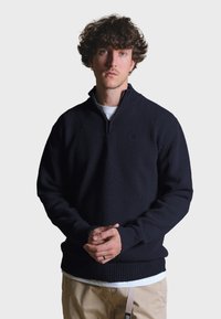 Tmavě námořnický pletený svetr s čtvrtím zipem, vroubkovaným lemem a manžetami, nošený přes bílou košili, s jednoduchým logem.