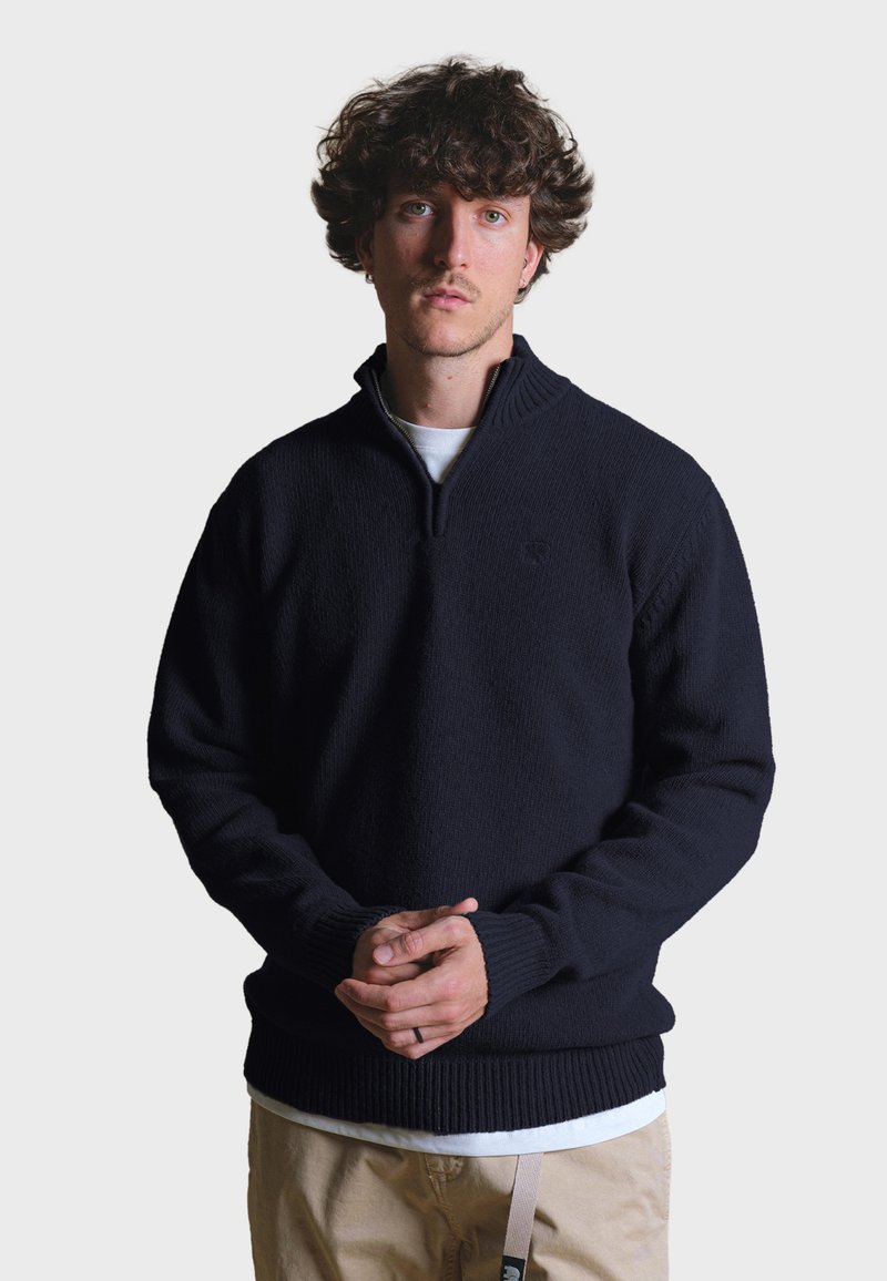 Tmavě námořnický pletený svetr s čtvrtím zipem, vroubkovaným lemem a manžetami, nošený přes bílou košili, s jednoduchým logem.