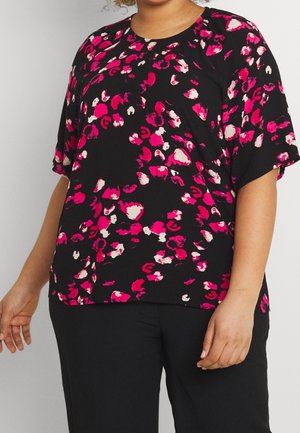 Vrouw gekleed in een zwart shirt met korte mouwen met een roze en witte abstracte bloemenprint en een zwarte broek, zichtbaar van schouders tot heupen.