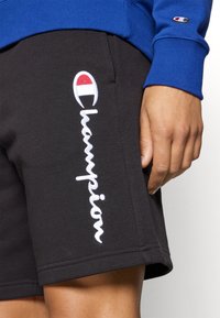 Pantaloni corti neri in cotone con il logo "Champion" ricamato di bianco sul lato, abbinati a un maglione blu con un logo sulla manica.