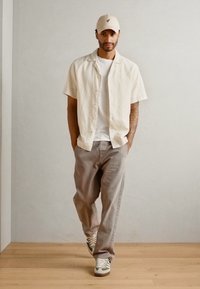 Ležérní outfit obsahující bílou košili s krátkým rukávem, šedé kalhoty a tenisky s zelenými akcenty, doplněný béžovou čepicí a hladkou texturou.