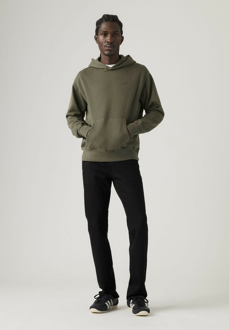 Junger Mann steht mit den Händen in den Taschen, trägt einen olivgrünen Kapuzenpullover, schwarze Hose und schwarze Sneakers vor einem schlichten Hintergrund.