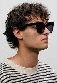 Černé obdélníkové sluneční brýle s tmavými skly, s jemným logem na stranici, které má na sobě osoba s kudrnatými vlasy, oblečená ve pruhovaném svetru.