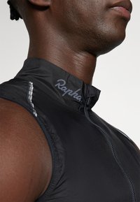 Maillot de cyclisme noir en tissu léger et lisse. Il possède un col haut et une fermeture éclair partielle avec un détail de bande réfléchissante.
