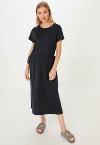 Schwarzes Kurzarmkleid mit elastischem Bund und seitlichen Schlitzen, aus weichem Stoff, kombiniert mit grauen Slippern.