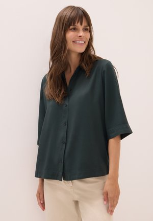 Lachende vrouw met lang bruin haar draagt een donkergroen overhemd met knopen en brede mouwen, en lichte beige broek, staande tegen een witte muur.