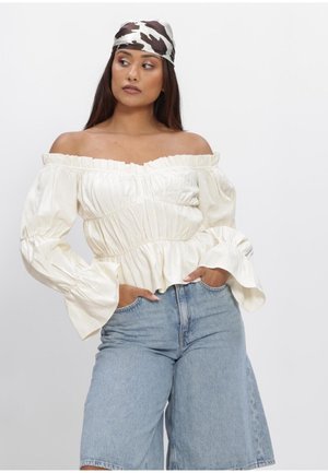Vrouw met een crèmekleurige off-shoulder blouse met plooien, lichtblauwe denim shorts met hoge taille en een hoofddoek met koeienprint, tegen een effen achtergrond.