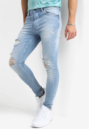 Mężczyzna w jasnoniebieskich podartych obcisłych dżinsach i białych trampkach, stojący na tle białego tła.