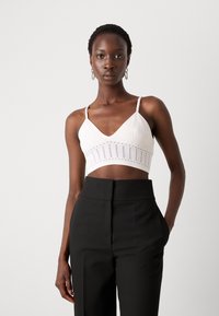 Weißes, kurz geschnittenes Bralette mit Spitze, V-Ausschnitt und dünnen Trägern. Kombiniert mit hochgeschnittenen, schwarzen, taillierten Hosen.